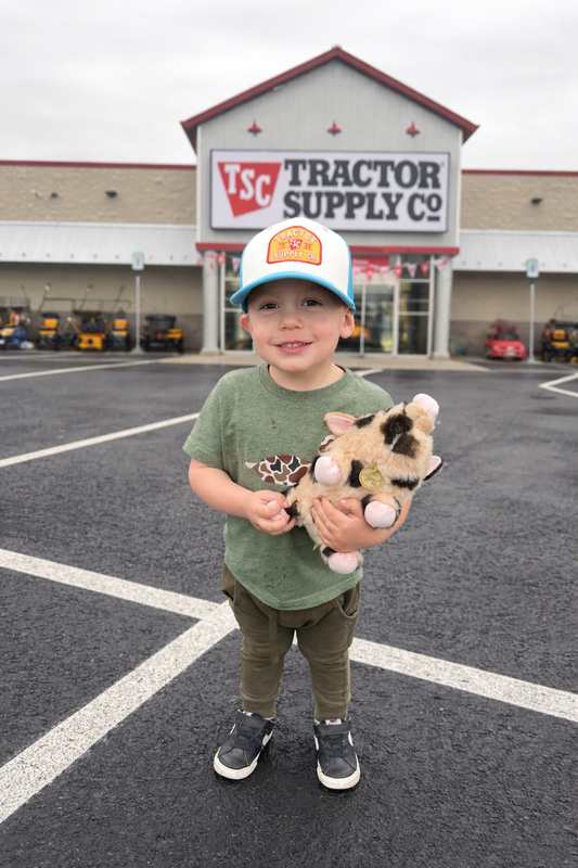 Toddler boy holding a plush pig toy standing in front of Tractor Supply Co. store wearing a green camo duck tee, matching green joggers and trucker hat - farm lifestyle kids clothing by Little Sprouts Outfitters