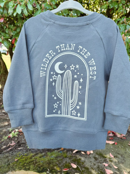 Blue sweatshirt with 'Wilder Than The West' design on a hanger against a natural background.