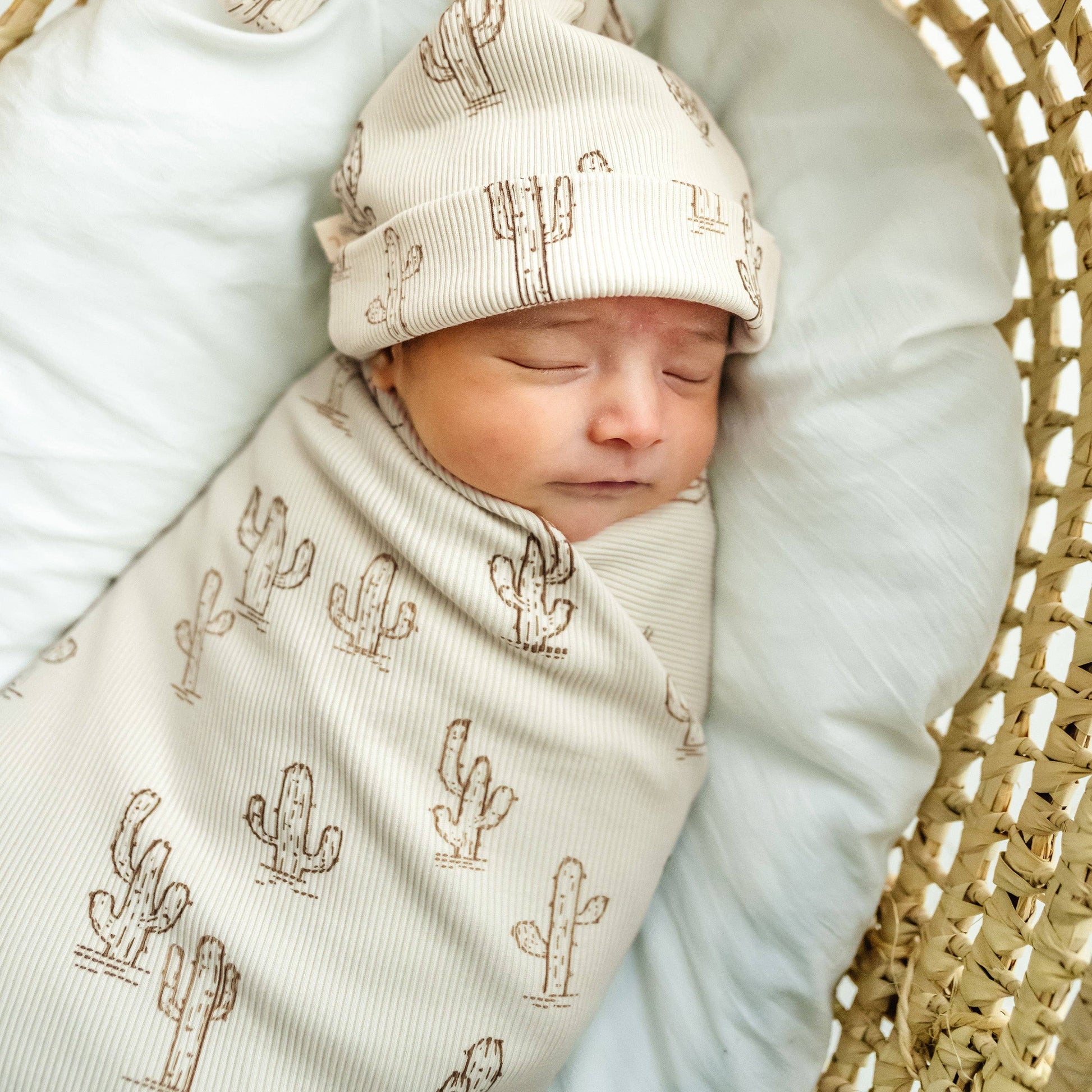 Newborn baby swaddled in a blanket with cactus pattern, wearing a matching hat, on a light blue cushion.