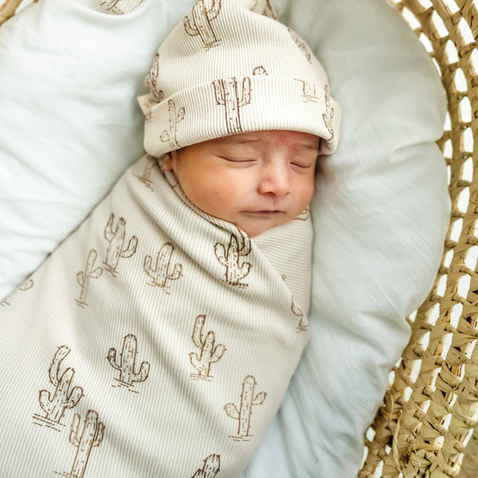 Newborn baby swaddled in a blanket with cactus pattern, wearing a matching hat, on a light blue cushion.