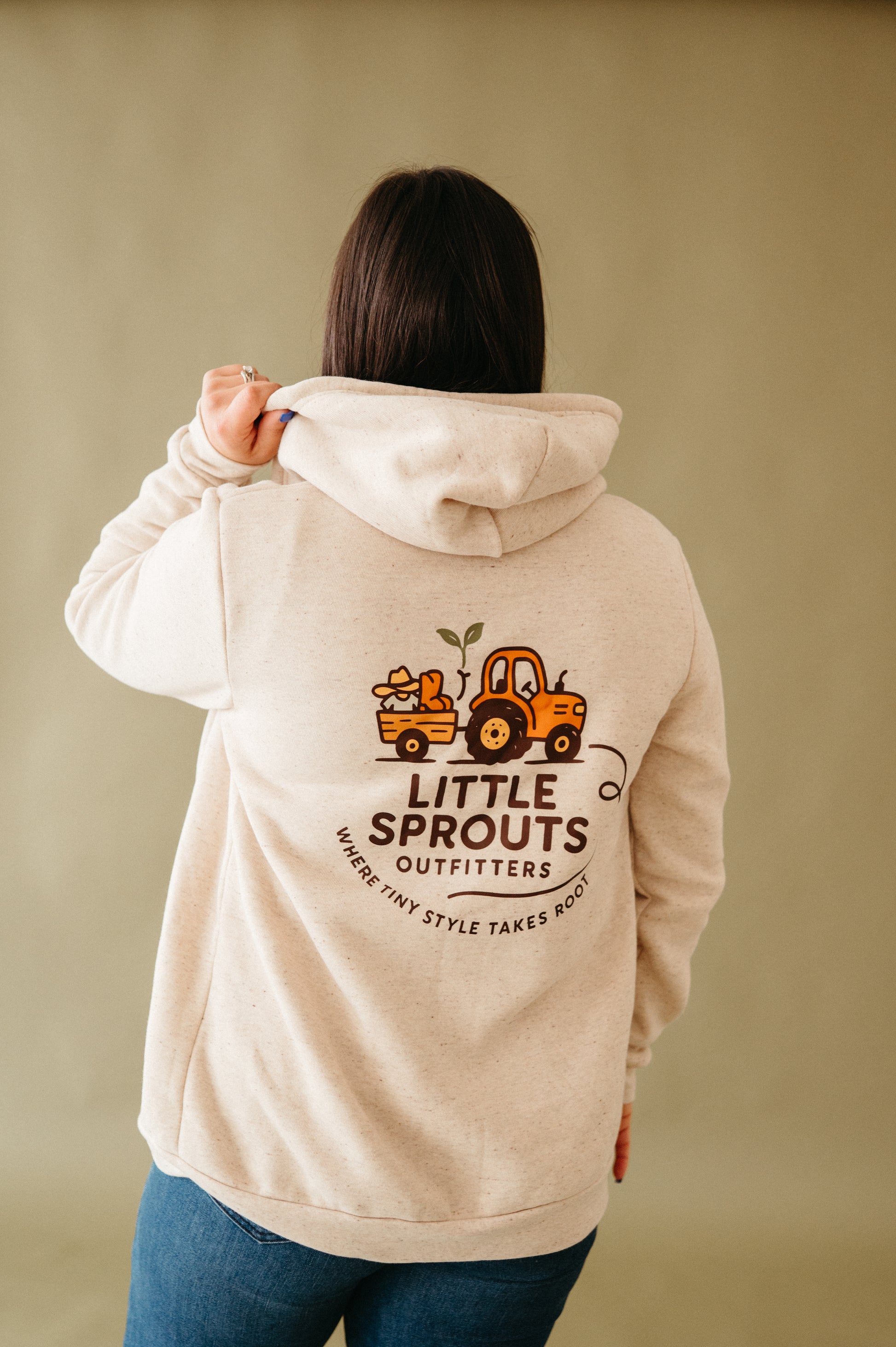 Person wearing a beige hoodie with 'Little Sprouts Outfitters' logo on the back against a beige background